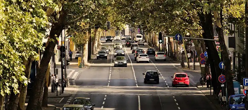 Das Bild zeigt eine Straße mit Autos und Fußgängern. Am Rand stehen grüne Bäume.